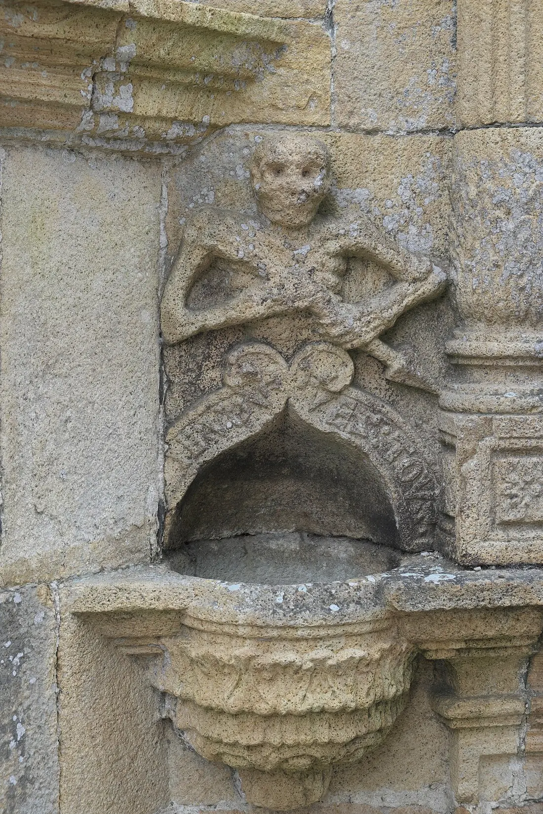 Squelette sculpté dans la pierre, au-dessus d'un bénitier, sur la façade d'un ossuaire